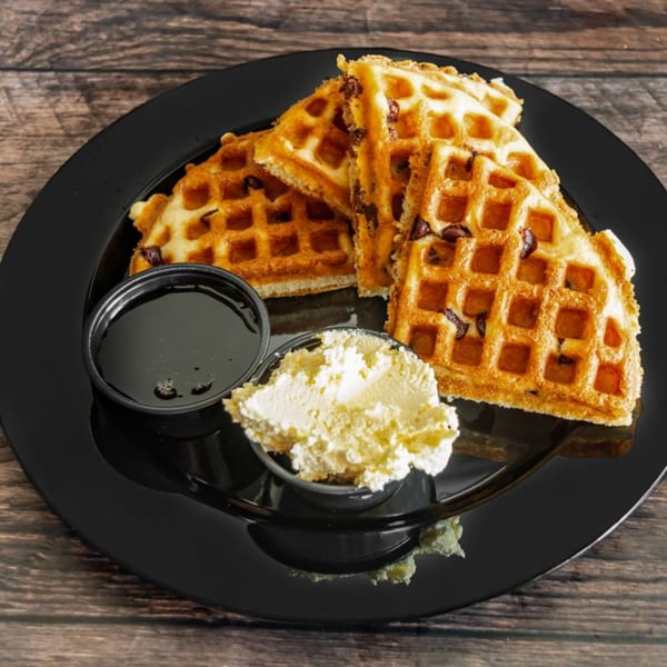 Golden waffles with chocolate chips, syrup, and whipped cream on a black plate.