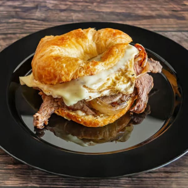 Savory croissant sandwich with roast beef, melted cheese, and caramelized onions on a black plate.