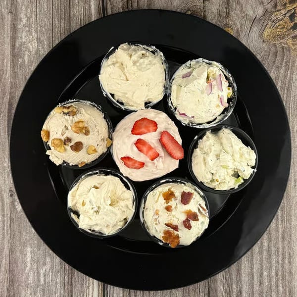 Seven small bowls of various cream cheese flavors, including strawberry, on a black plate.