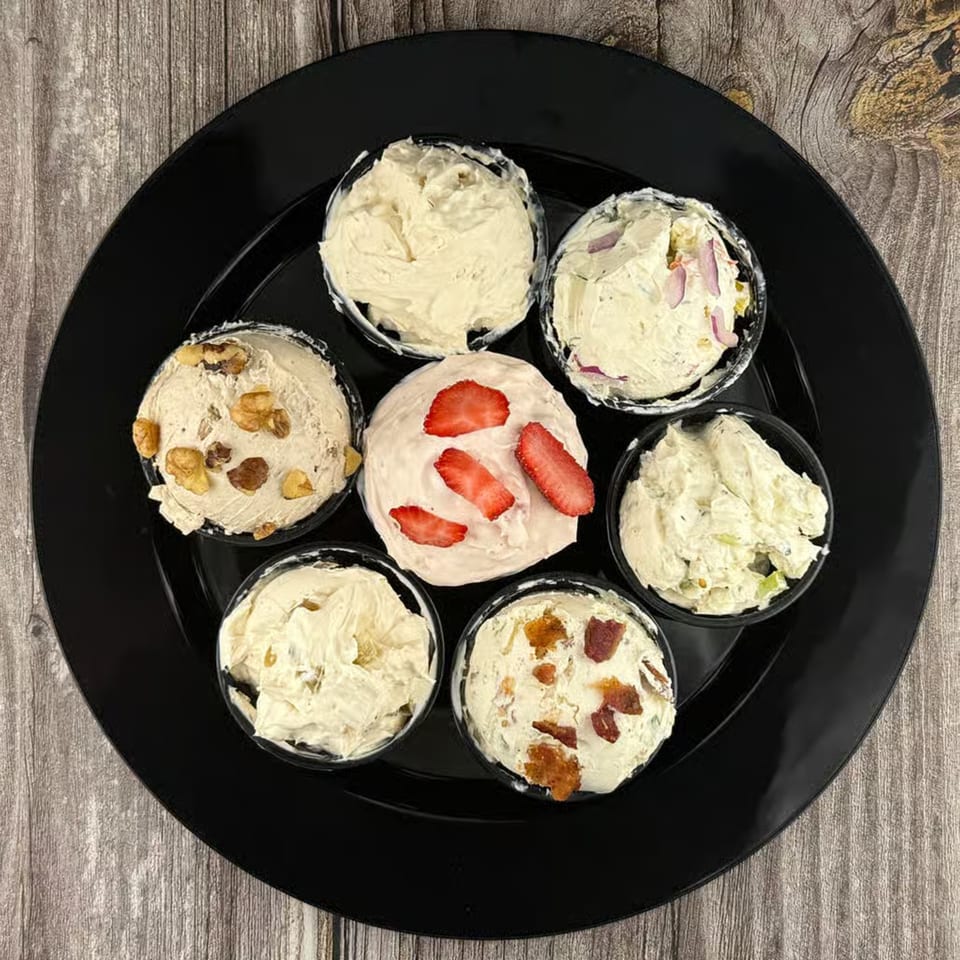 Seven small bowls of various cream cheese flavors, including strawberry, on a black plate.