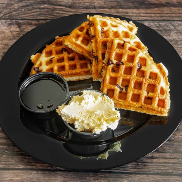Golden waffles with chocolate chips, syrup, and whipped cream on a black plate.
