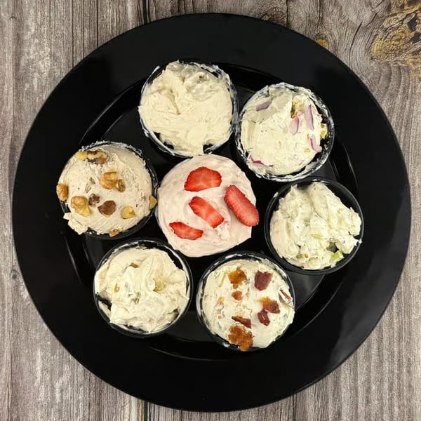 Seven small cups of various cream cheese flavors, including strawberry, on a black plate.
