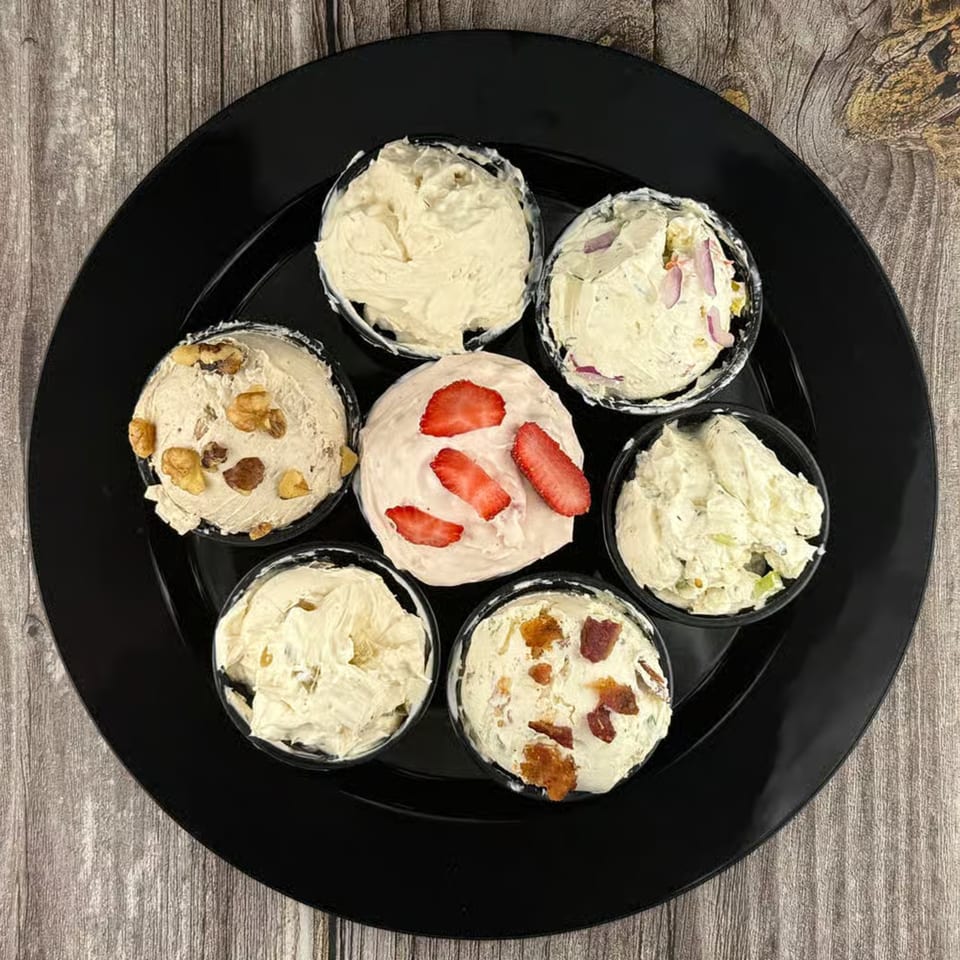 Seven small cups of various cream cheese flavors, including strawberry, on a black plate.